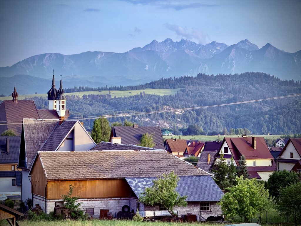Picturesque view of a Polish village with a church, houses, and barns in the foreground, framed by the dramatic Tatra Mountains in the background under a clear blue sky.