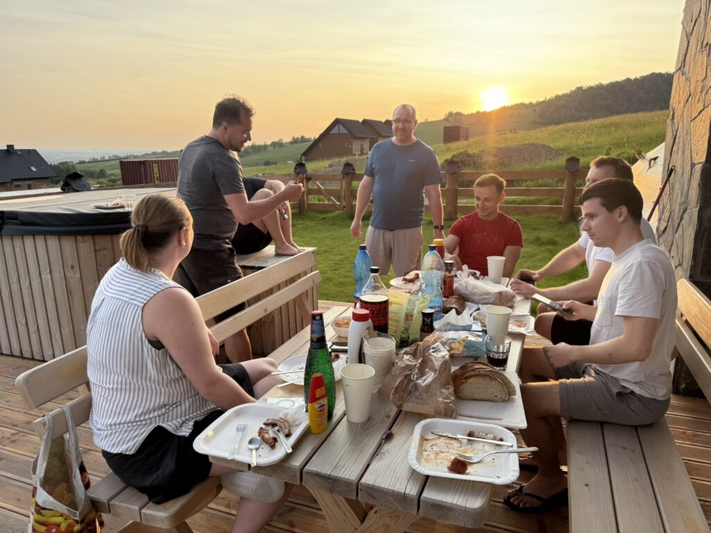 Group of people enjoying an outdoor dinner on a wooden terrace during sunset in the countryside, with a hot tub nearby and food, drinks, and bread spread across the table.