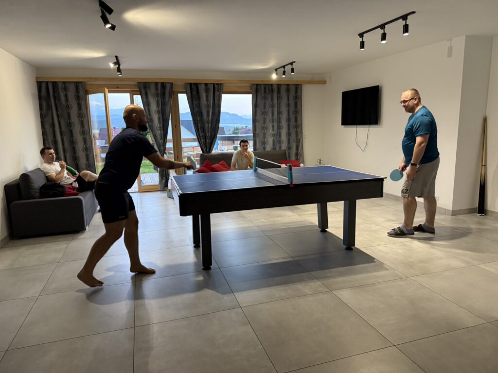 Two men playing table tennis in a modern living room with large windows and a mountain view, while two others relax on couches watching the game.