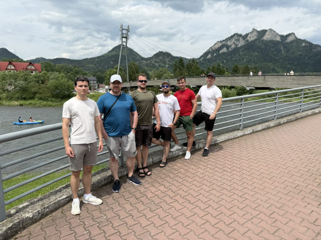 Six men stand side by side on a riverside walkway with a suspension bridge and the distinctive rocky peaks of the Pieniny mountains in the background. Behind them, a group in a blue kayak paddles on the river, and several people are visible walking across the bridge. The mood is casual and relaxed, with a mix of neutral and sporty clothing.