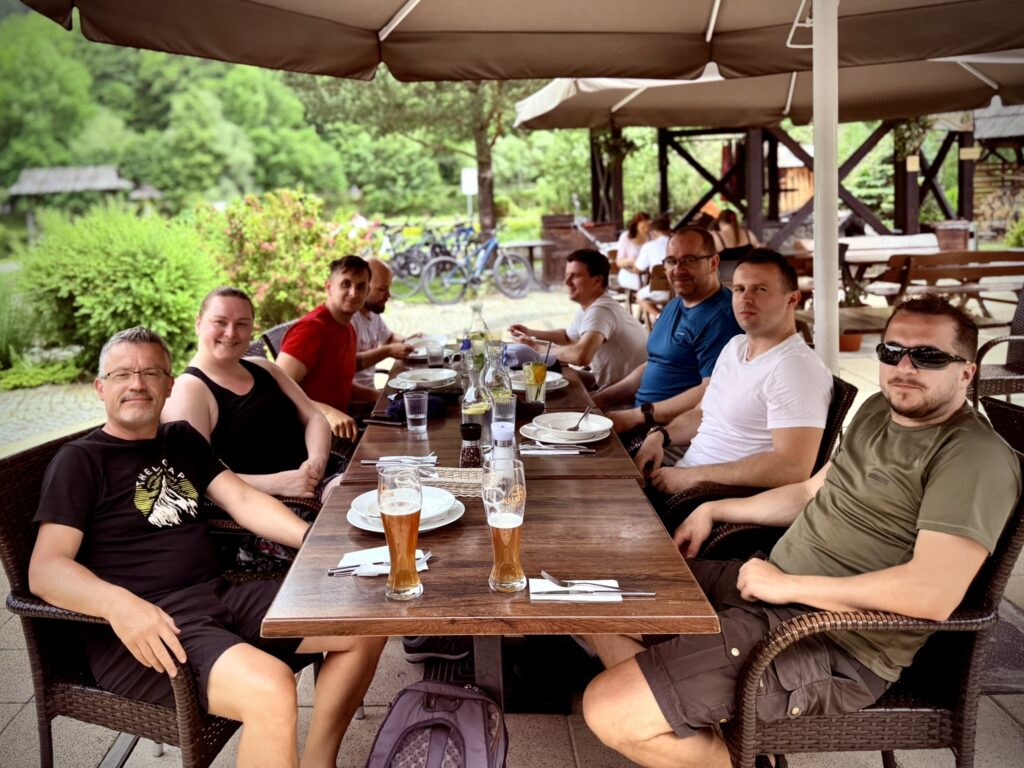 A group of nine people sits around a long wooden table at an outdoor restaurant, enjoying a relaxed moment before their meal. The table is set with plates, utensils, and drinks, including a few glasses of beer. In the background, there are bicycles, lush greenery, and wooden structures under large umbrellas. The mood is friendly and casual.