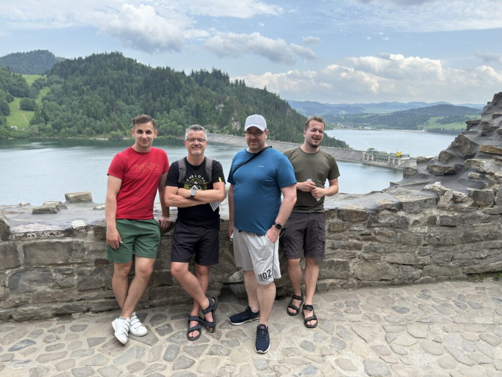 Four men pose together on a stone castle terrace overlooking a scenic reservoir. Behind them, a forested hillside and a dam stretch out under a partly cloudy sky. The man in red and the one in black stand casually in front, while the man in blue and the one in olive green lean in slightly, all smiling or relaxed in the moment.