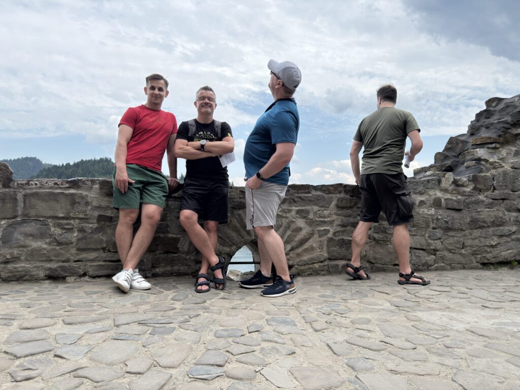 Four men stand on a stone terrace of a historic castle. Two lean casually against the wall; one in red looks straight ahead, the second in black crosses his arms. The man in a blue shirt looks up into the sky, while the one in olive green gazes down toward the ground beyond the wall. Rolling hills and cloudy skies complete the scenic backdrop.