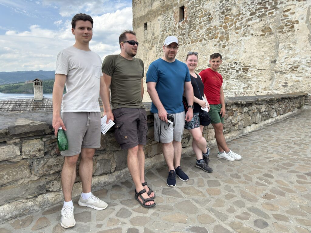 Five people stand in a row, leaning against a stone wall with a historic fortress wall behind them. The group is casually dressed for warm weather, enjoying a relaxed moment during their visit. A lake and distant hills are partially visible in the background.