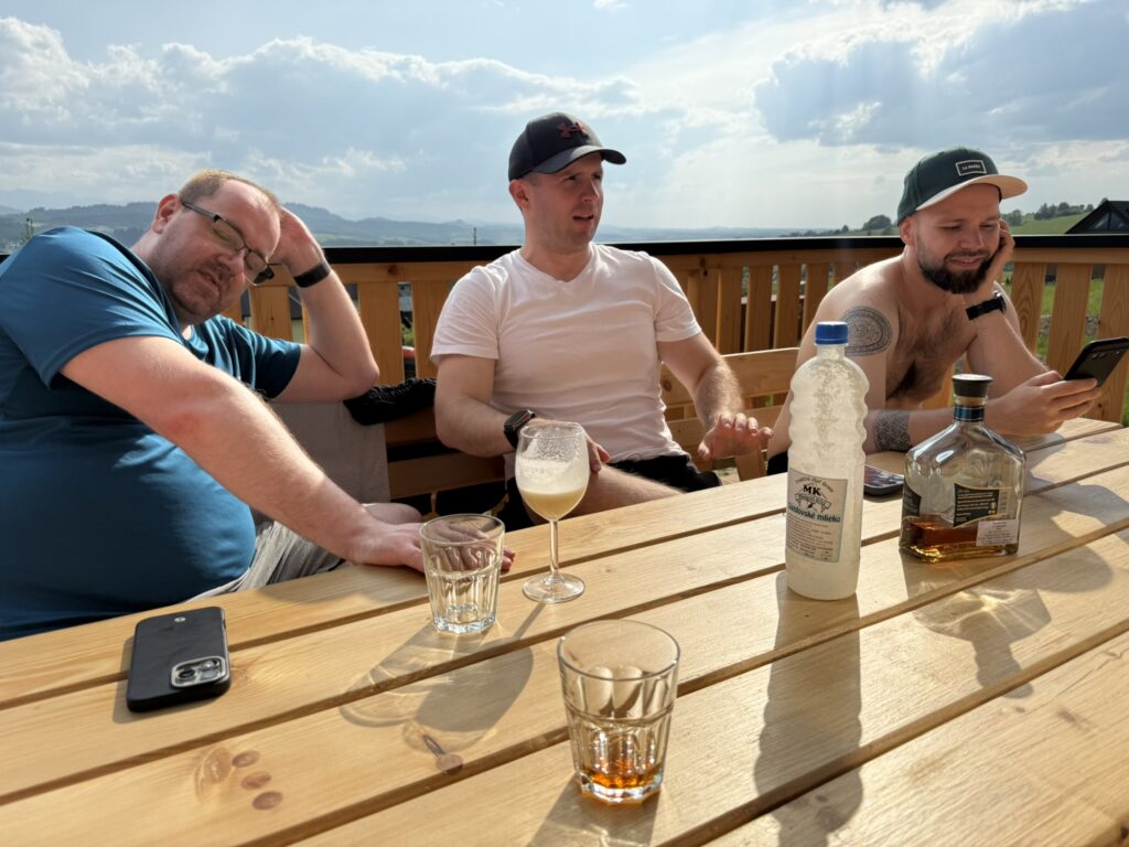 Three men are sitting at a wooden terrace table with drinks in front of them. One man is leaning his head on his hand, the second is talking, and the third is looking at a phone and smiling. There are bottles, including one of milk and one of alcohol, on the table. The background shows hills and cloudy sky.