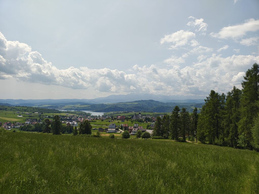 A peaceful countryside stretches out under a bright sky, with a small town nestled among rolling green hills. In the distance, a lake glimmers, and the silhouettes of mountain peaks rise beyond the horizon. Tall trees frame the right side, adding a natural border to this idyllic scene.