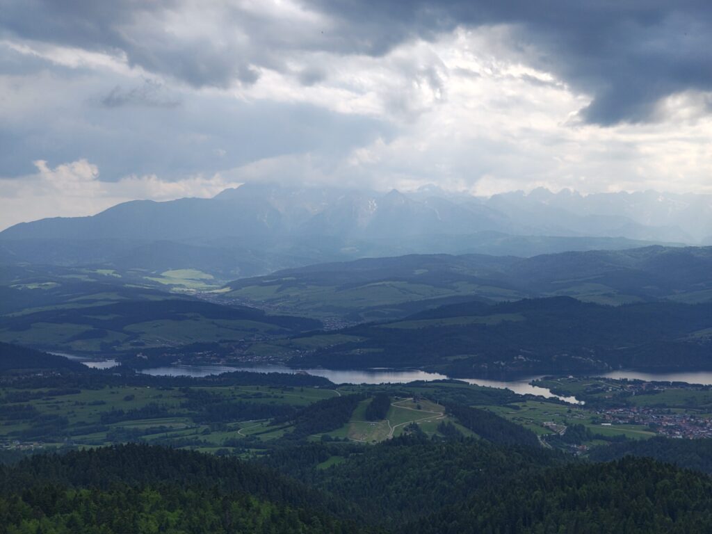 A dramatic landscape unfolds beneath a stormy sky, where shafts of sunlight break through thick clouds to reveal a patchwork of green hills, winding rivers, and distant mountain silhouettes.