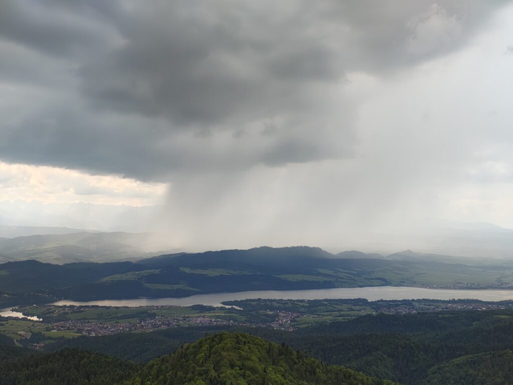 A dramatic rain shower descends from heavy clouds over the landscape, creating a striking contrast between light and shadow across the hills and lakes below.