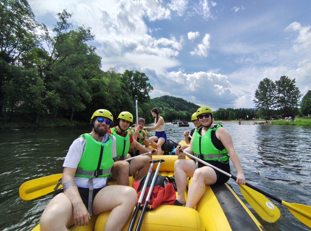 A cheerful group of people rafting on a calm river, wearing yellow helmets and bright green life vests, enjoying a sunny day surrounded by forested hills and blue skies.