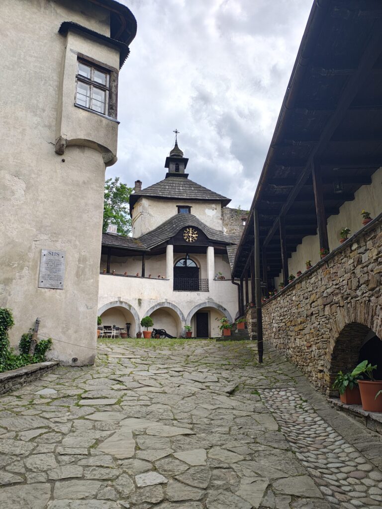 A courtyard of a historic castle with stone pavement, arched passages, flower pots, and a clock tower with a wooden shingle roof under a cloudy sky.