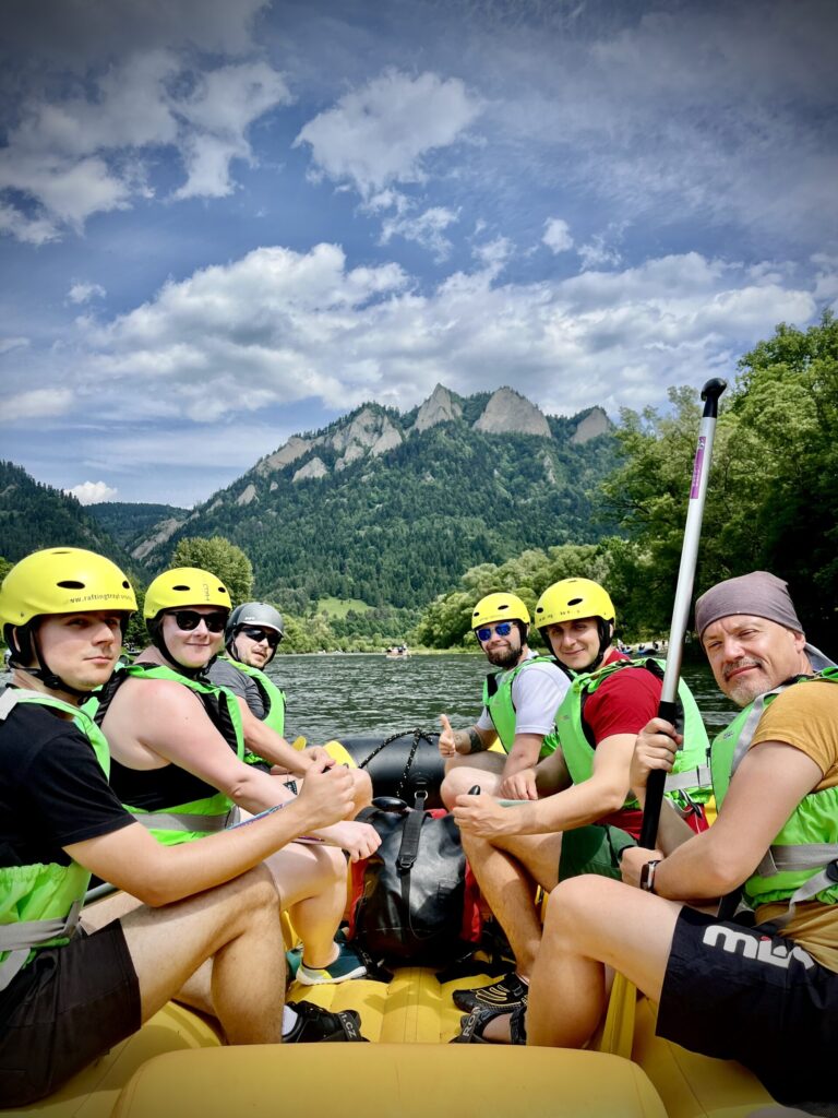 A cheerful rafting group paddles down a calm river, framed by lush green trees and the striking limestone peaks of the Pieniny Mountains in the background. The vibrant yellow helmets and green life vests pop against the natural scenery, capturing the thrill of adventure in a picturesque setting.