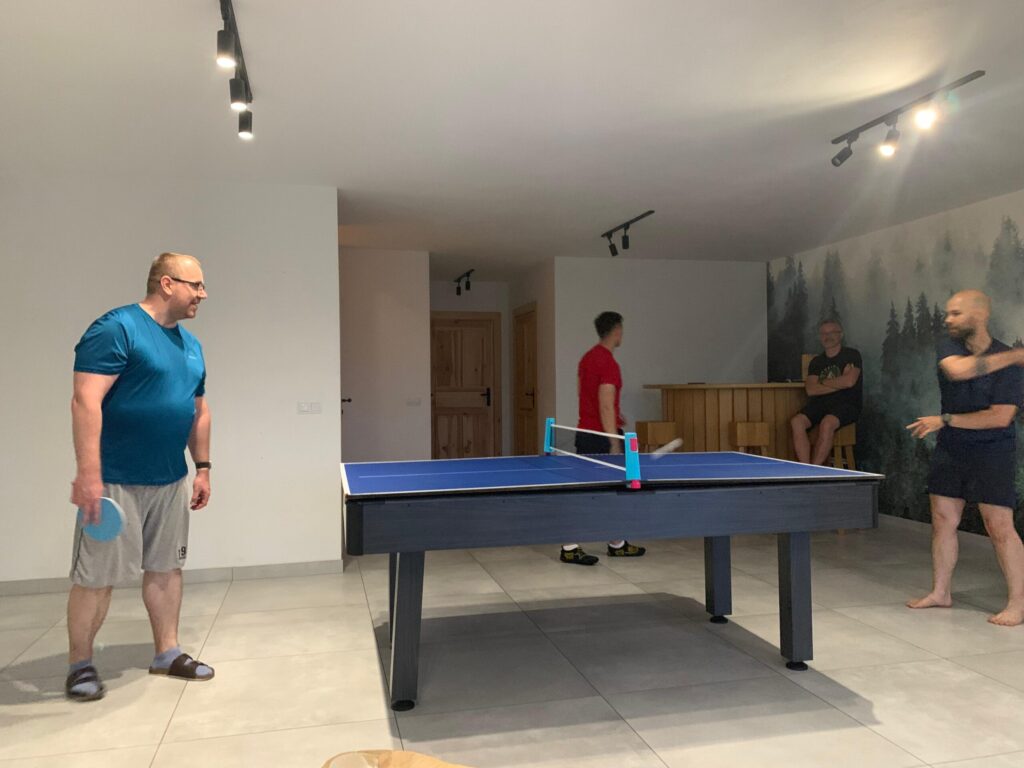 Four men playing table tennis and watching inside a modern room with a forest-themed wall mural and a wooden bar.
