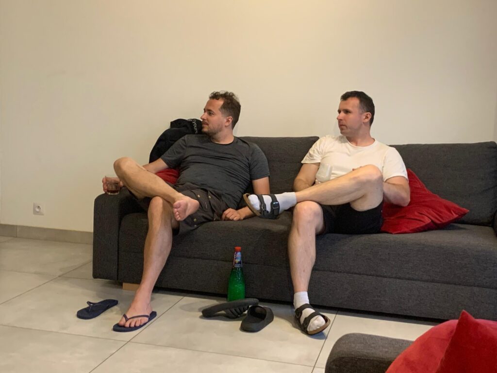 Two men sitting on a grey couch with red pillows, watching a ping pong game while holding drinks and relaxing.