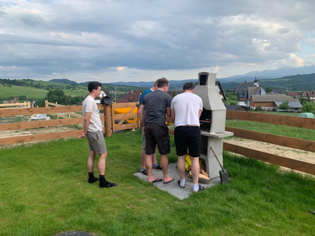 Four men grilling food at a concrete barbecue in a scenic Polish village, surrounded by rolling hills and rooftops, during a team-building event.