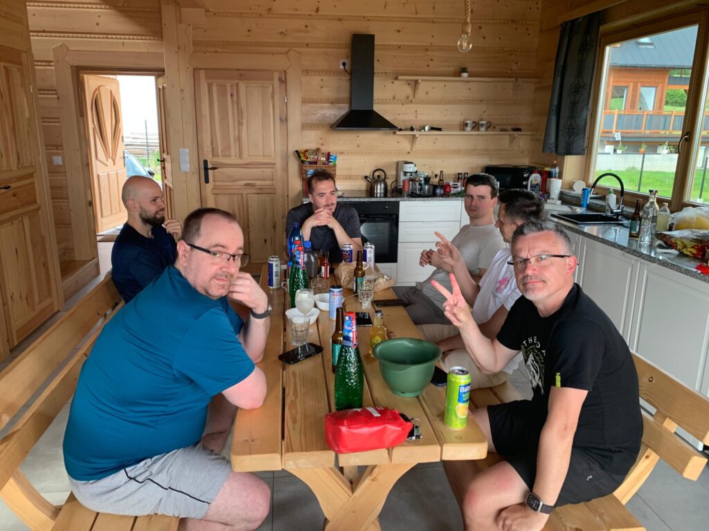 Group of men sitting around a wooden table inside a cozy log cabin kitchen, enjoying drinks and snacks during a teambuilding event.