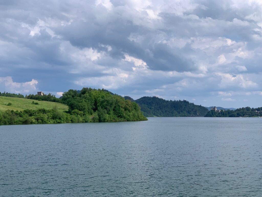 A peaceful lake surrounded by green hills and forests under a cloudy sky, with two distant castles visible on opposite sides of the landscape.