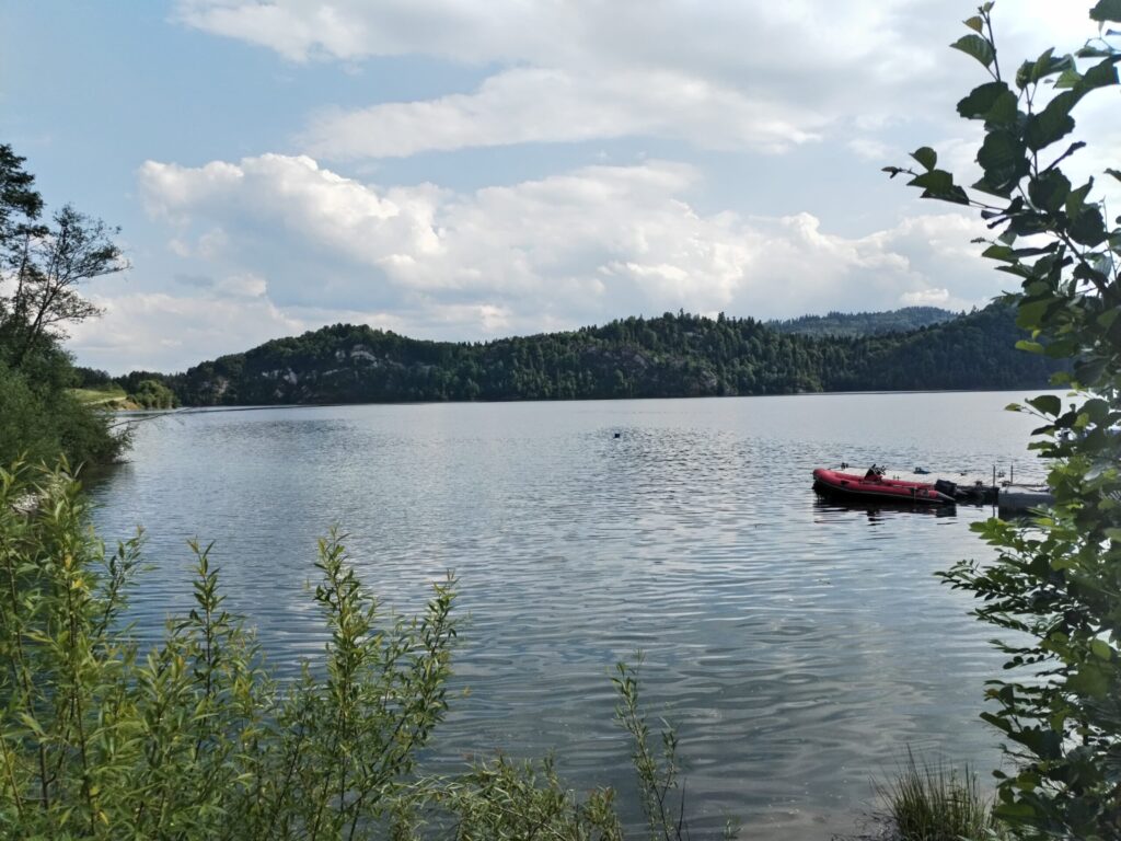 A calm lake surrounded by green forested hills under a partly cloudy sky, with a small red inflatable boat docked near the shore and bushes framing the foreground.